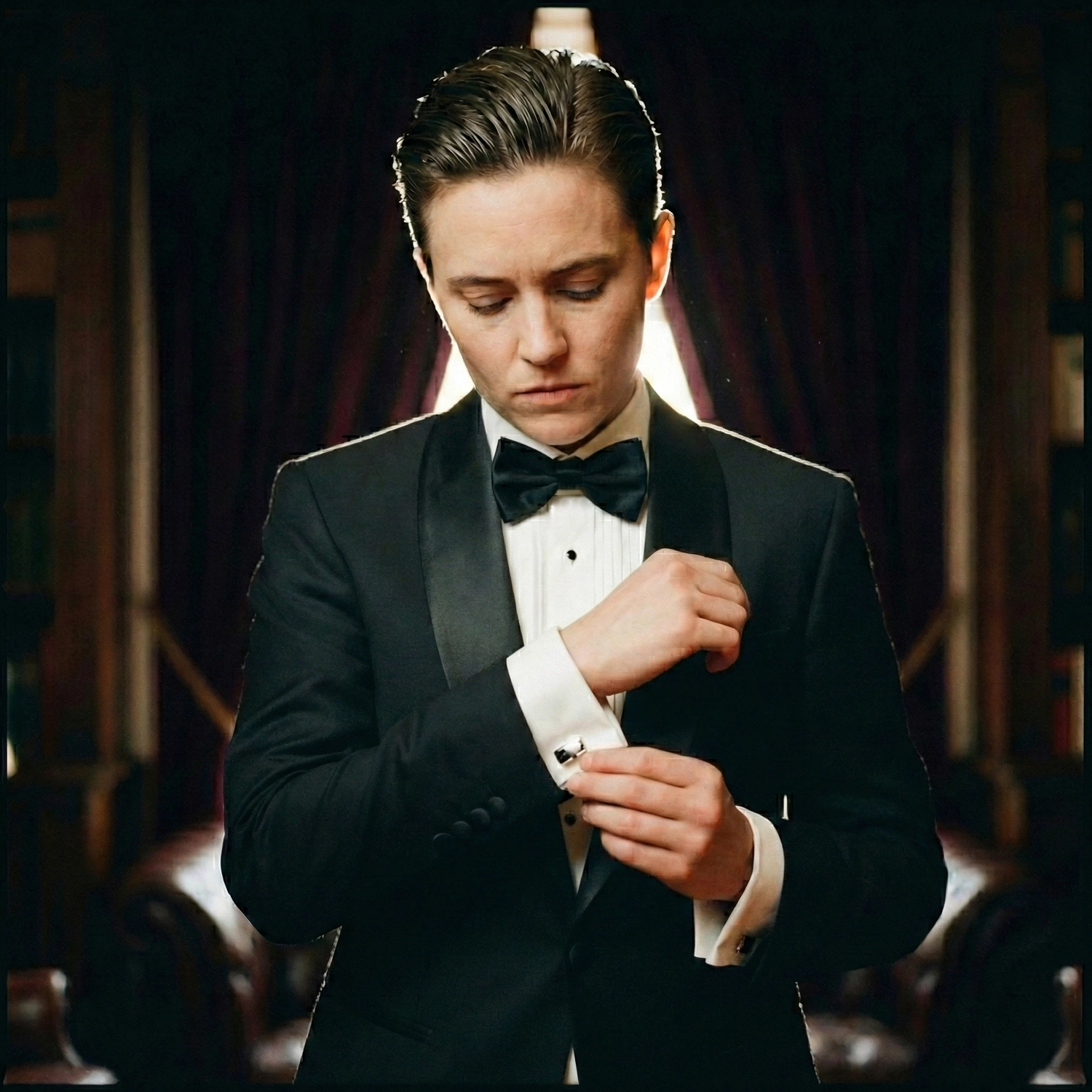 Person in a black tuxedo adjusting cufflinks, showcasing elegant androgynous wedding accessories like a black bow tie in a room with dark curtains and leather chairs.