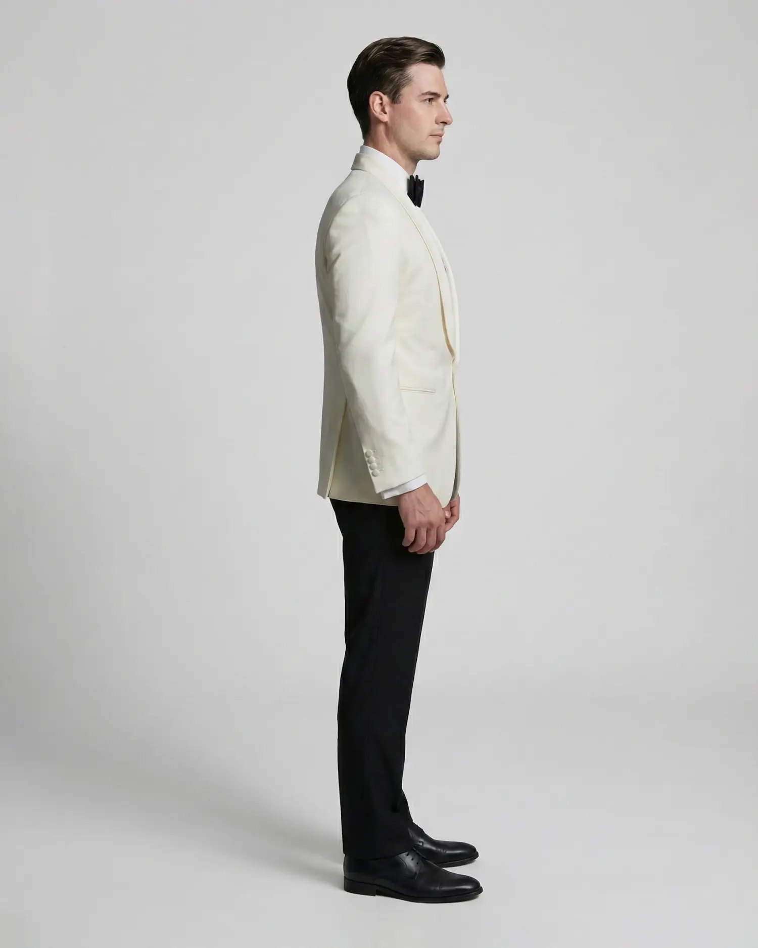 Man in white tuxedo with black bow tie standing in profile against a gray background.