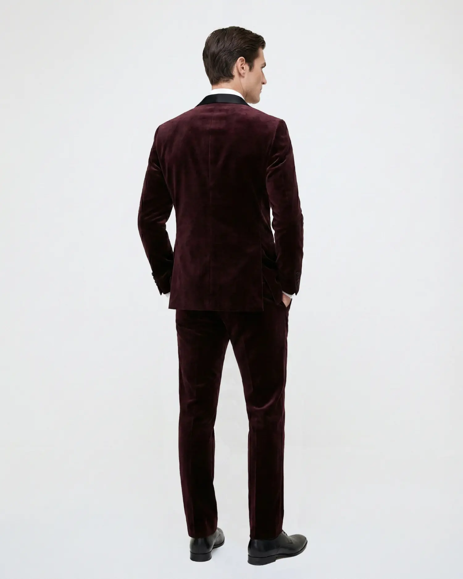 Man in a burgundy velvet suit standing with back turned, showcasing the luxurious fabric against a plain background.