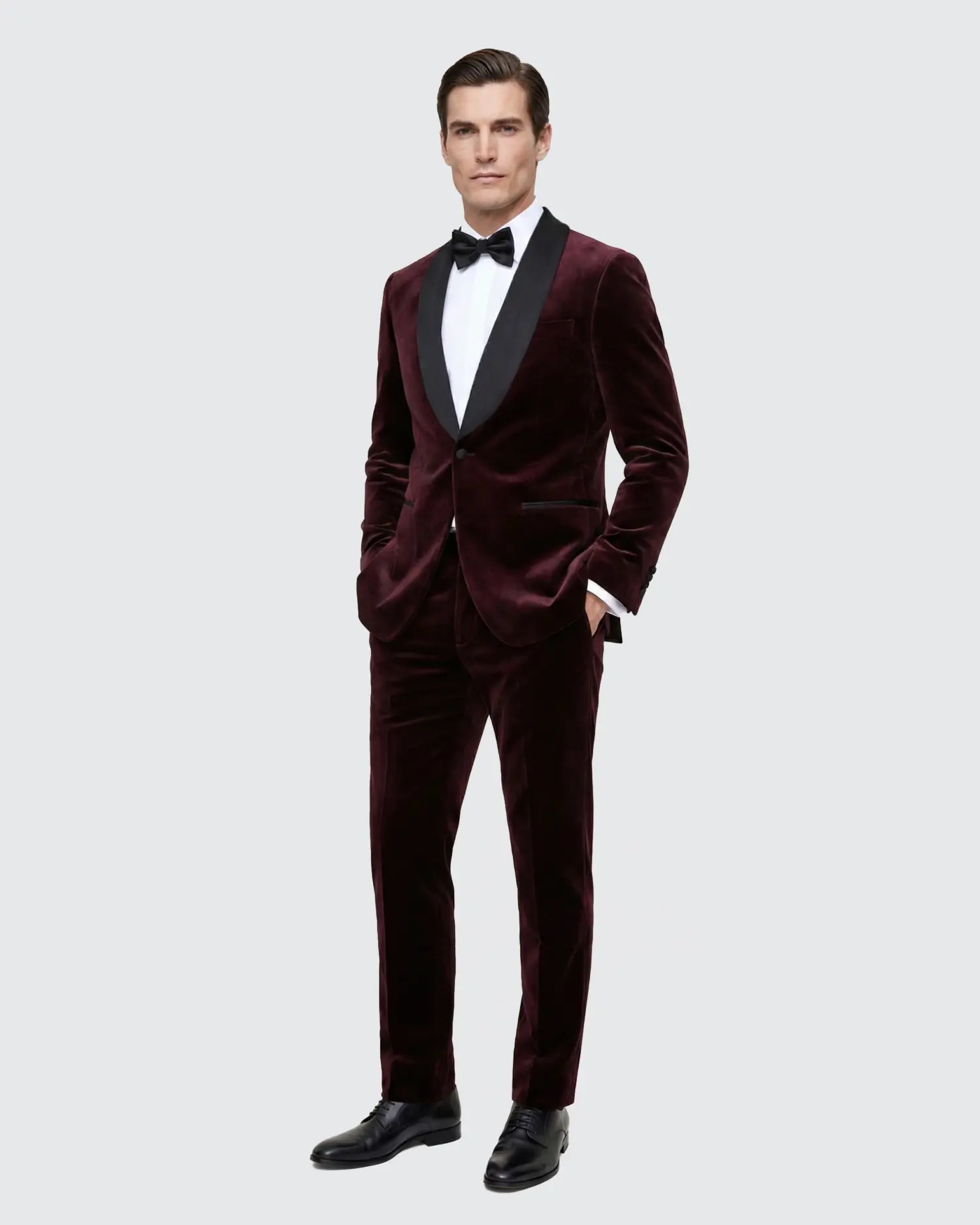 Man in a burgundy velvet tuxedo with black lapels, white shirt, and bow tie, standing against a light gray background.
