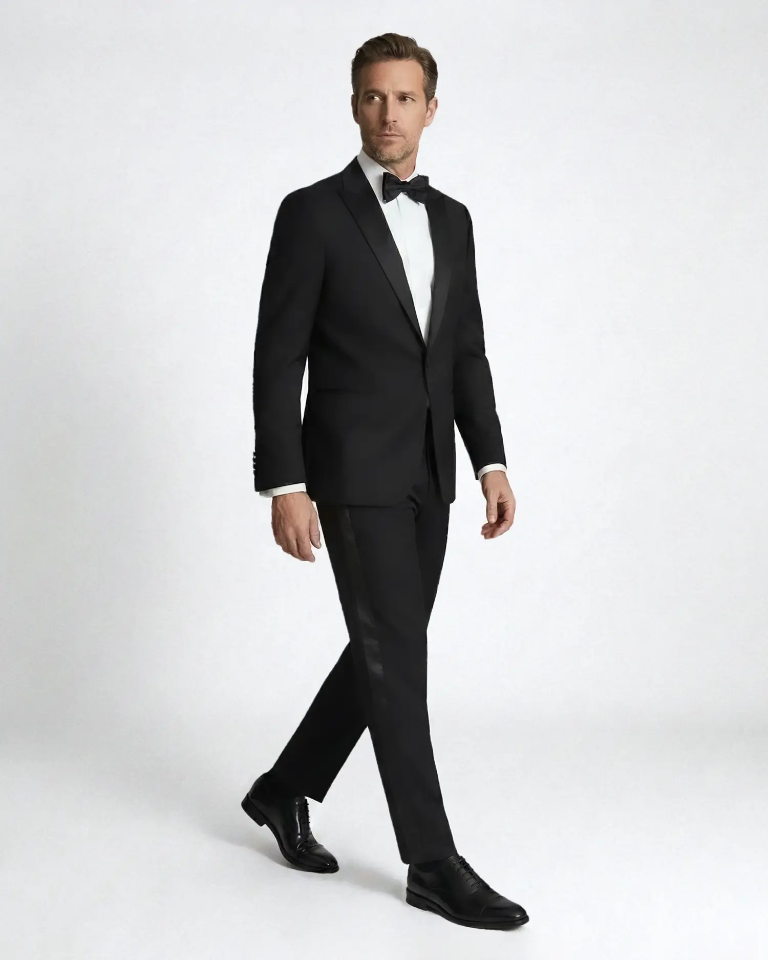 Man in a formal black tuxedo with a bow tie, walking confidently against a neutral background.