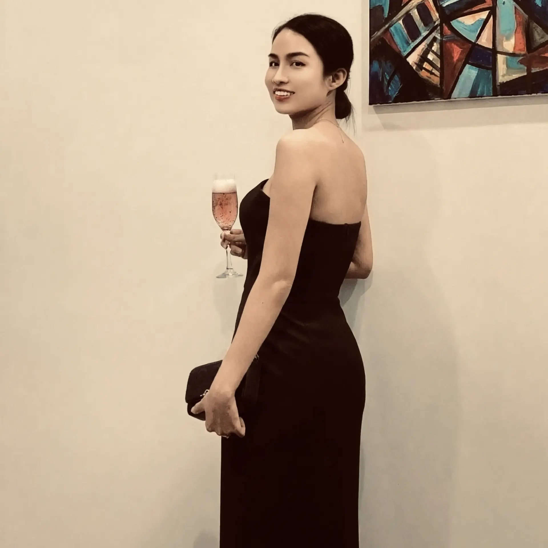 Woman in elegant black dress holding a glass of champagne stands by abstract painting on wall.