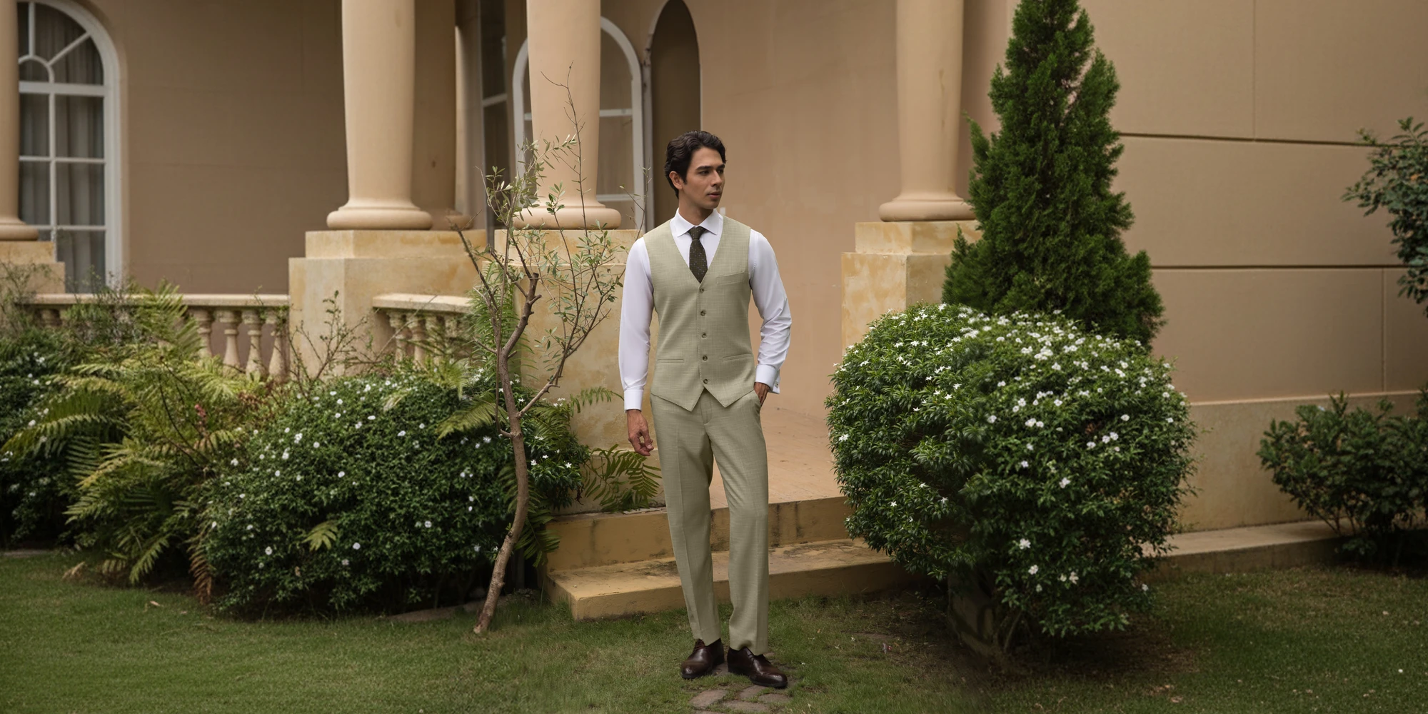 Man in elegant beige suit, standing confidently in a garden setting with architectural backdrop.