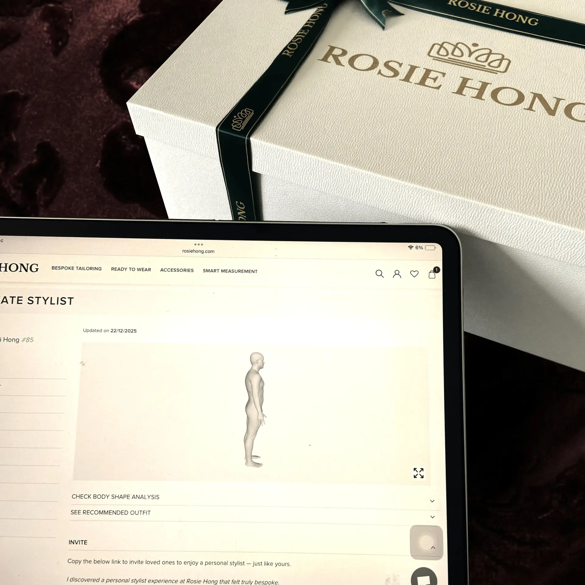 Tablet displaying 3D model next to Rosie Hong gift box with logo and ribbon on brown surface.
