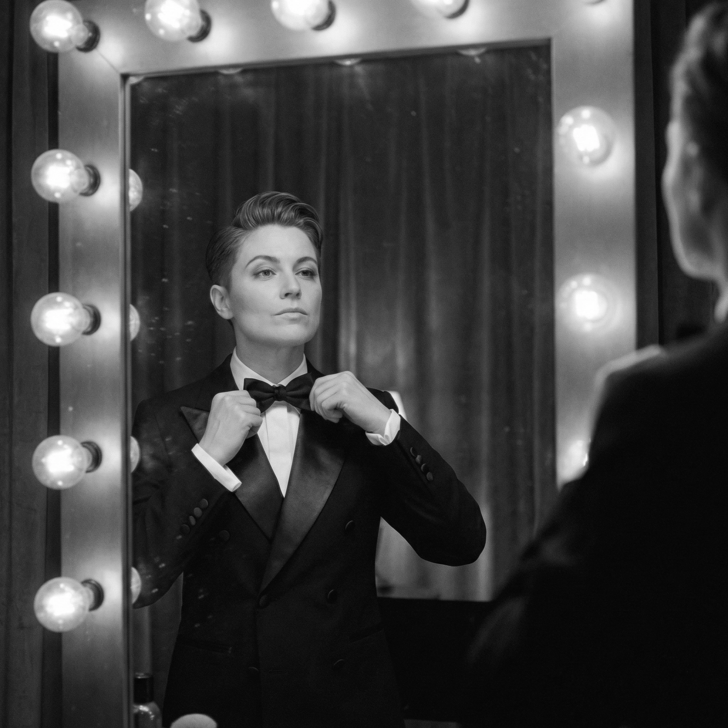 Person adjusts bow tie in front of a vanity mirror with lights, dressed in a formal suit, confident expression. Black and white.
