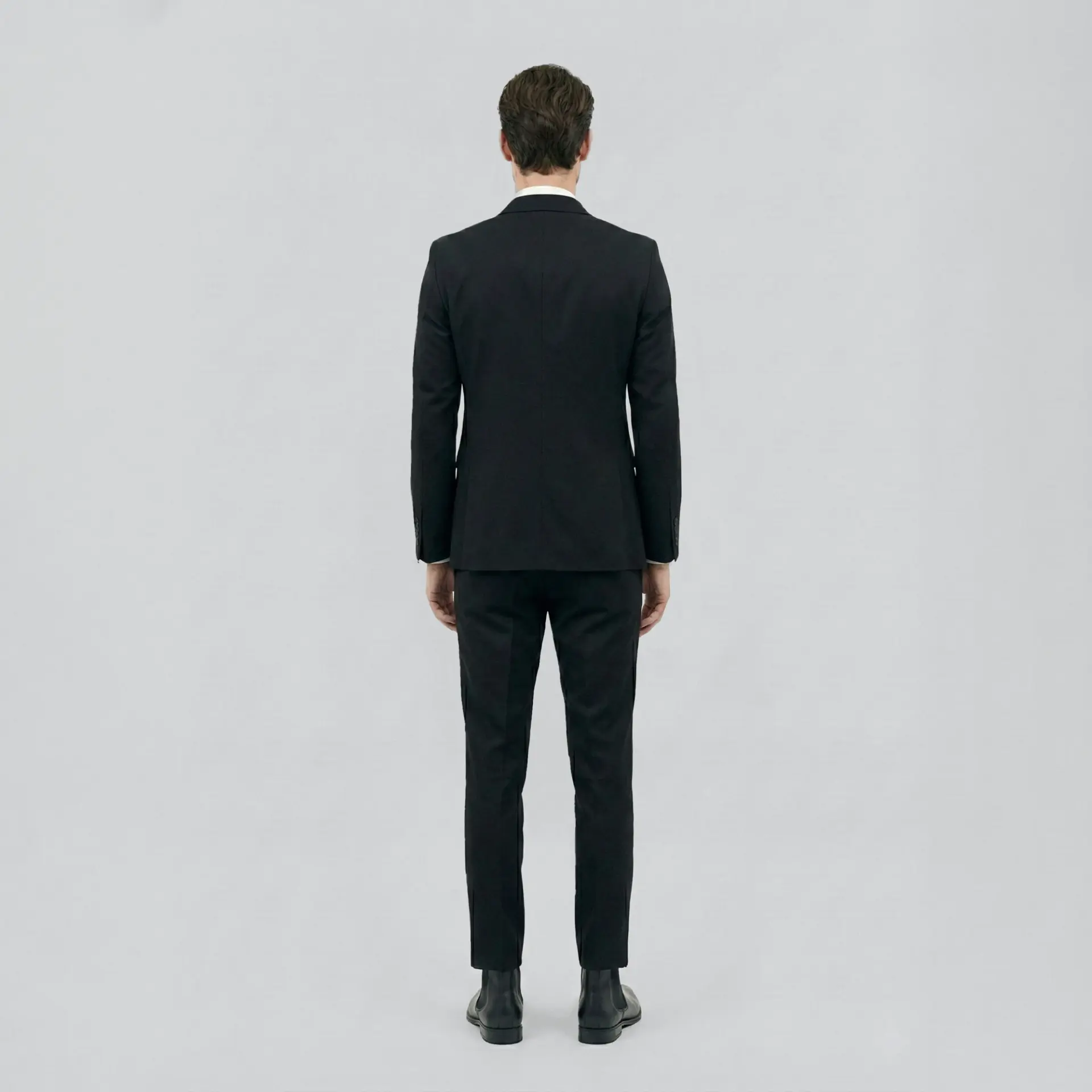 Man in a black suit standing with back facing, on a light grey background.