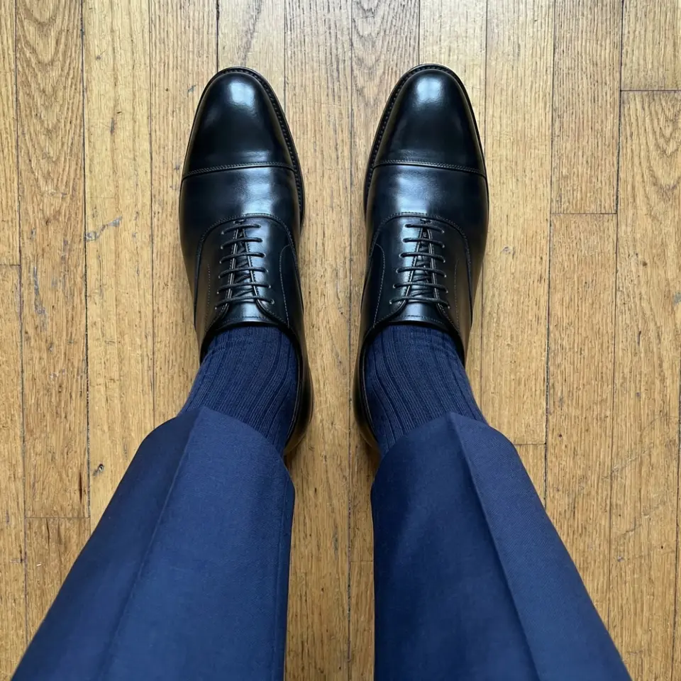 Black leather dress shoes with navy socks and trousers on a wooden floor.