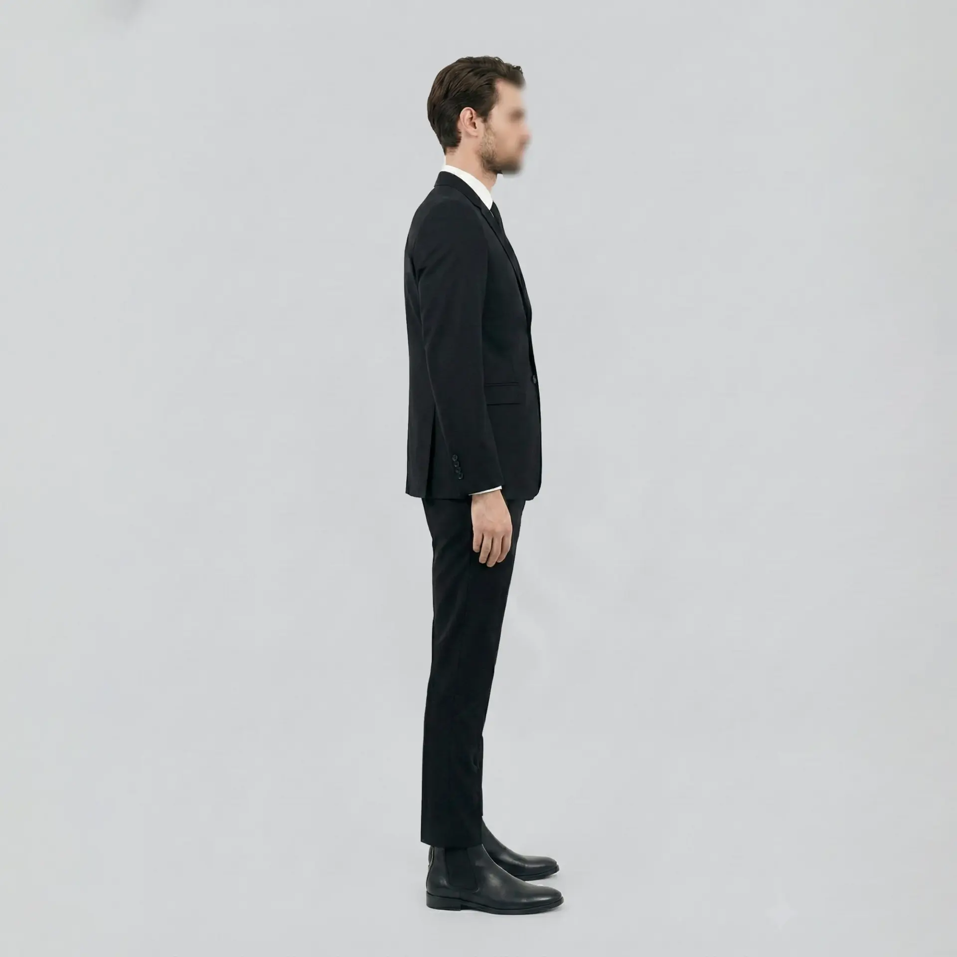 Man in black suit standing sideways on a plain background, showcasing professional attire.