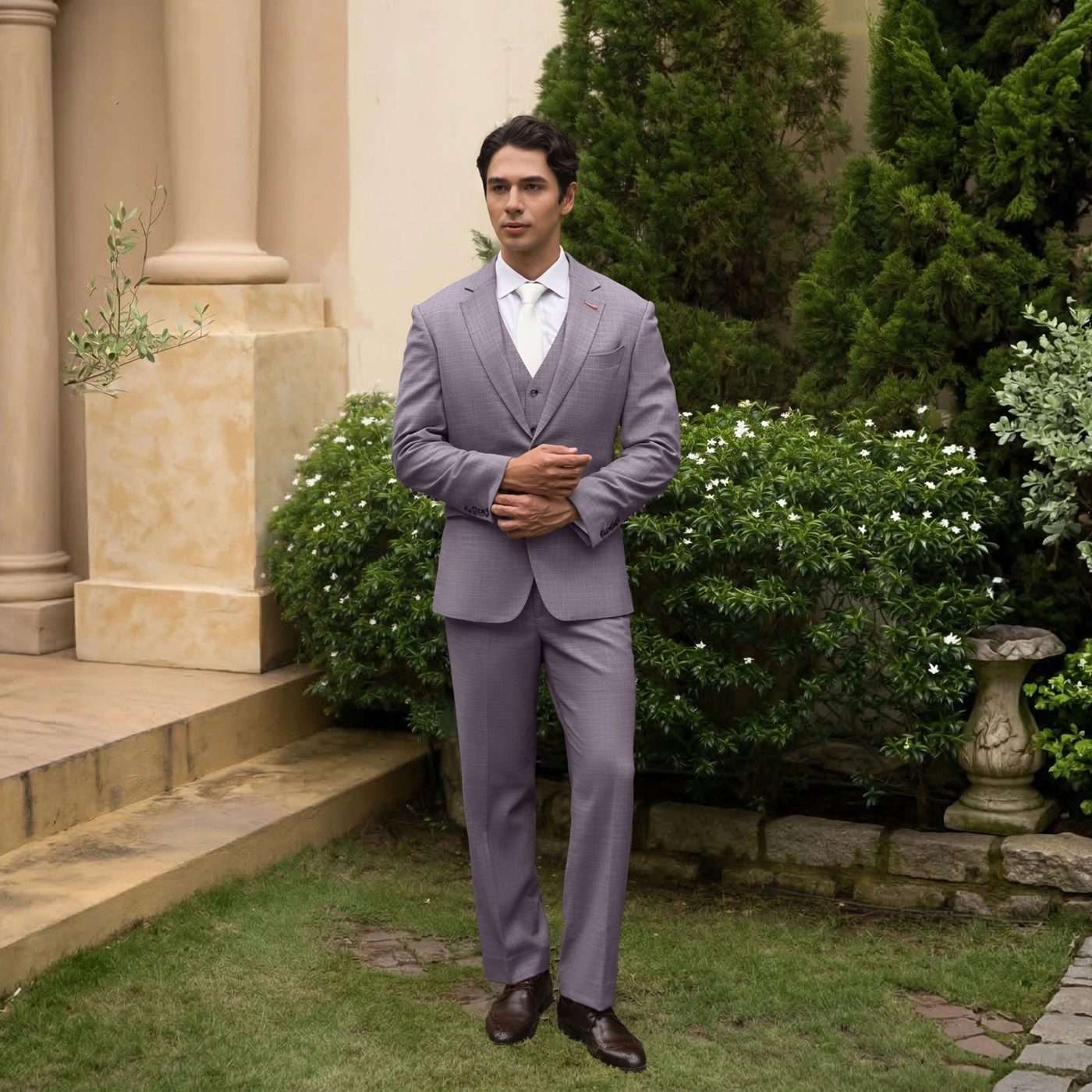 Man in a tailored suit stands in a garden setting, showcasing elegant formal wear against lush greenery.